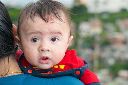 beautiful latin baby peeking his head over his mother's shoulder, big baby with amazed face, blue overalls and red shirt looking back. with his mouth open and big cheeks. concept of motherhood.の写真素材