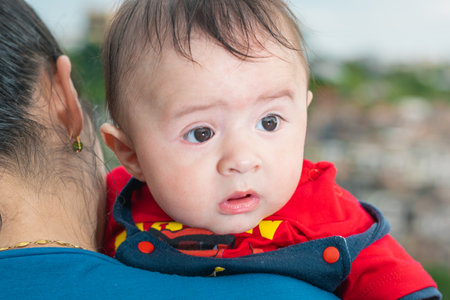 beautiful latin baby peeking his head over his mother's shoulder, big baby with curious look, blue overalls and red shirt looking to his left. with his mouth open and big cheeks. concept of motherhoodの写真素材