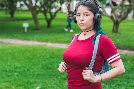 beautiful college woman with blue hair, red dress and blue backpack, listening to music with her wireless headphones in a park. green background. concept of freedom.の写真素材