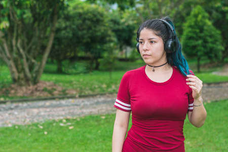 beautiful college woman with blue hair and red dress, listening to music with her wireless headphones in a park, stroking her blue hair while looking to the right. relaxation conceptの写真素材