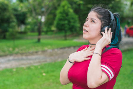 beautiful college woman with blue hair and red dress, listening to music with her wireless headphones in a park, excited and sentimental girl listening to songs with one hand on her chestの写真素材