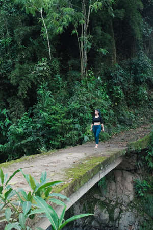 beautiful latin woman crossing a cement bridge covered with green moss in the middle of the Colombian jungle, observing nature feeling free and without fear of the void (height).の写真素材