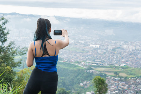 latina woman on the top of a mountain shooting a video of the city of Pereira-Colombia, with her mobile phone in horizoltal. girl hiking in the colombian mountains.の写真素材