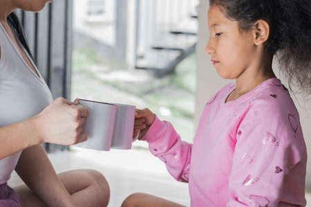beautiful brown-skinned latina girl with her mother toasting with white cups. the girl looks bored while remembering that her father is absent. concept of motherhood.の写真素材