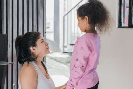 beautiful latina mother sitting on the floor while looking her brown-skinned daughter in the eyes, the girl is standing next to her mother who advises her and is proud of her. family conceptの写真素材