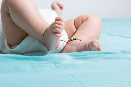 beautiful and tender feet of a two months old baby, lying on a bed with blue sheets, on one of her feet she has an ankle bracelet and is wearing a white romper suit.の写真素材