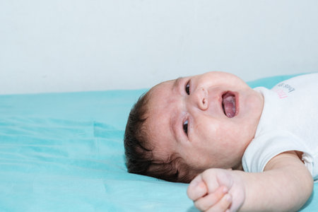 beautiful latina baby crying loudly because she is hungry, lying on a bed with blue sheets. baby with angry gesture and her mouth open screaming. concept of anger.の写真素材
