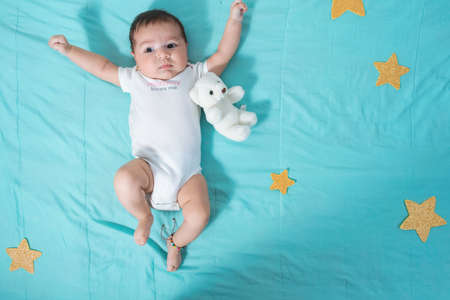 beautiful latin baby girl, two months old, next to a small teddy bear, raising her hands and looking distracted, surrounded by stars and a sea blue savannah background.の写真素材