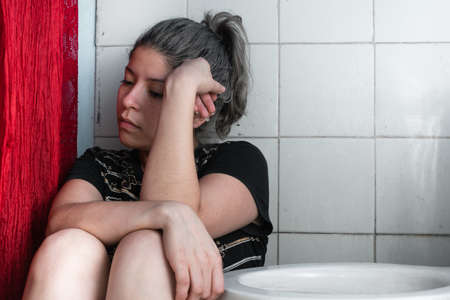 beautiful latina woman, with her hair painted gray, sitting on the side of the toilet, very pensive and depressed because of so many problems in her life. bored girl with one hand on her head.の写真素材