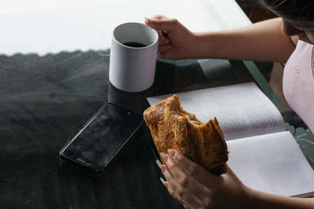 beautiful latina woman holding in her left hand a sandwich she is eating before going to the office. girl reading a book while drinking coffee in a white mug. her cell phone is on the table.の写真素材