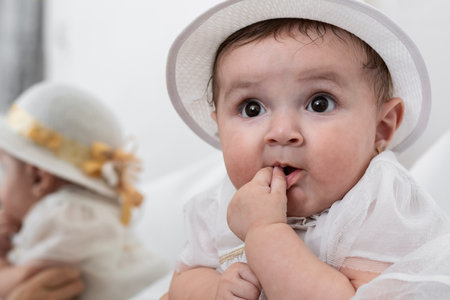 close-up of a beautiful white-skinned latina baby girl with a white dress and hat, looking with curiosity and a little smile at her mother, with a hand on her mouth. baby at her christening.の写真素材