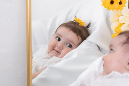 beautiful face of a latin baby girl, seen in detail in the reflection of a large mirror decorated with yellow and white flowers. little girl with a yellow flower on her head.の写真素材