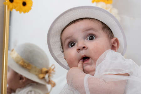 beautiful baby girl with a white hat, looking impressed at her mother while inserting the fingers of her left hand into her mouth. baptism of a baby girl in a catholic church.の写真素材