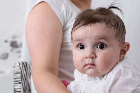 beautiful baby with big eyes, sitting on her mother's lap after receiving food. little girl looking curiously with her mother behind her. baby dressed in whiteの写真素材