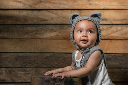 beautiful baby latina with brown skin, wearing overalls and a cap with mouse ears, knitted with the crochet technique. with orange wooden background and resting on a wooden chair.の写真素材