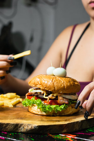 close-up of a latin girl with her hamburger and fries served on the wooden table, ready to start eating. woman with a potato in her hand about to put it in her mouth. street food.の写真素材