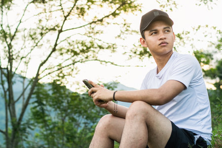 young latin man, sitting on the top of a mountain with his cell phone in his hand and a beautiful golden sunrise in the background, happily looking at the nature around him.の写真素材