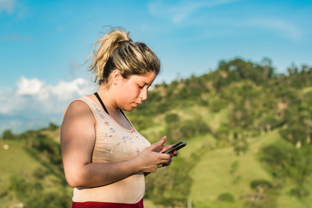 beautiful young latina woman, on top of a mountain with her cell phone in her hand, interacting on social networks, looking for information with the help of the satellite internet.の写真素材
