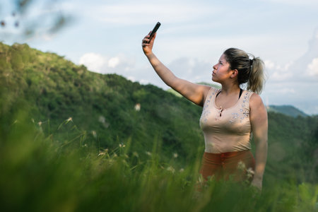 beautiful young latina woman, on top of a mountain looking for network coverage for her cell phone. girl looking for satellite signal, hiking on top of a hill. technology conceptの写真素材