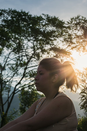 beautiful latin girl looking bored at the horizon with a beautiful sunset in the background, surrounded by trees on a summer day. concept of freedom and peace of mind.の写真素材