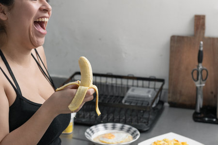 beautiful latina woman with a banana in her hand laughing out loud, girl eating a banana in her kitchenの写真素材