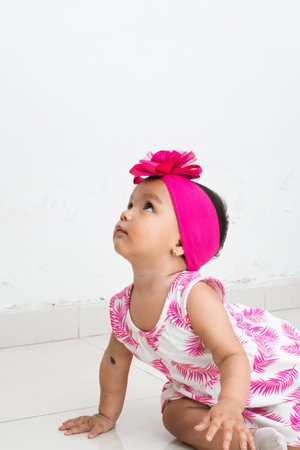 baby latina sitting on the floor looking upwards, with a bench dress with fuchsia leaves. white backgroundの写真素材