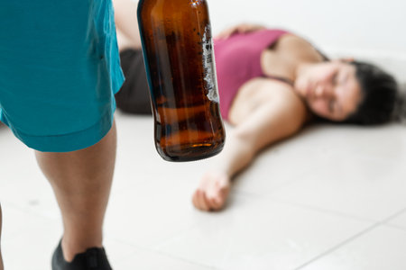 man with a beer bottle in his hand, leaving his wife lying on the floor after having beaten her. domestic violence and abuse of womenの写真素材