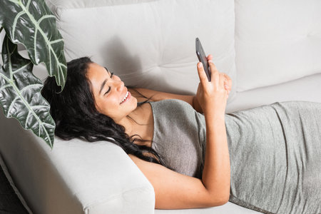 millennial brunette woman smiling while looking at her cell phone, lying on a white sofa spending her free time looking at her social networks.の写真素材