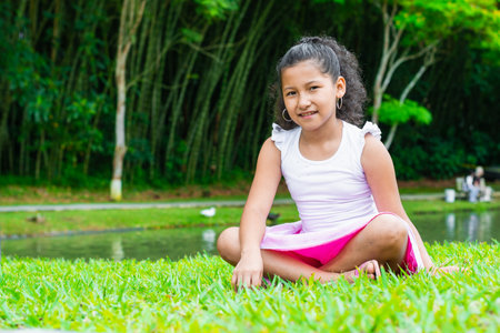 portrait of a little brunette girl, sitting on the grass next to a lake, smiling while looking straight aheadの写真素材