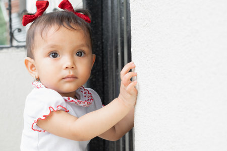beautiful baby brunette latina, standing next to the wall looking curiously, while holding her hands on the wall.の写真素材