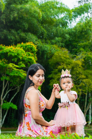 latina mother sitting on the grass looking at her beautiful brunette baby proudly, strolling through a green park with big trees.の写真素材