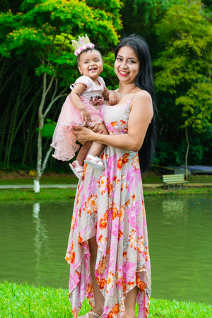 portrait of a Latina mother holding her little brown baby girl, standing by a lake in a park.の写真素材