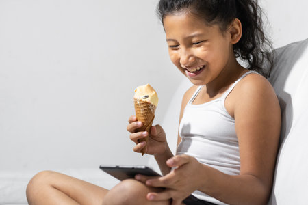 happy, smiling little brunette girl eating an ice cream while playing on her tablet or cell phone, sitting on the couch in her living room.の写真素材