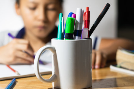 detailed view of a white cup full of pens and school supplies, in the background a little Latina student girl writing on her notebook.の写真素材