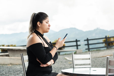 young latina woman, looking at her cell phone, while sitting in an outdoor restaurant, waitingの写真素材