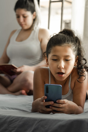 little latin girl, lying on the bed looking at her cell phone with an astonished face, her mother is...