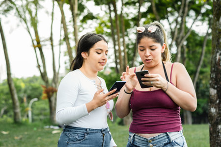 two latina girls looking at their cell phones in the park, taking a walk in the open airの写真素材