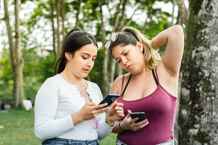 Latina women in the park, one with a look of discomfort at what she is looking at on her friend's cell phone.の写真素材