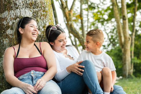couple of latin girls, sitting next to a tree in a park laughing loudly with their little sonの写真素材