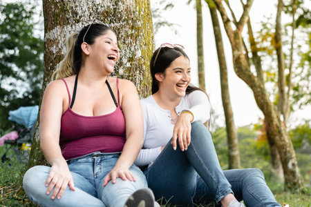 two friends sitting next to a tree, laughing loudly while looking to the sideの写真素材