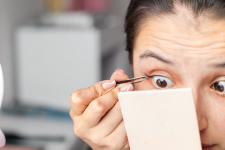 detailed shot of a latina woman using a manual eyebrow tweezer to apply false eyelashesの写真素材