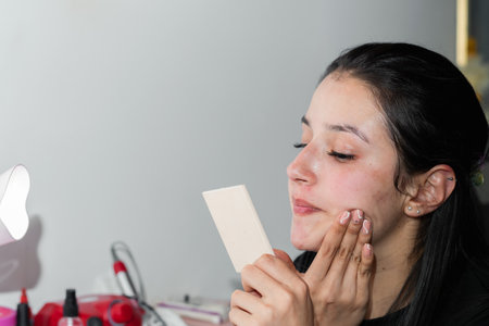 young latina woman applying a skin moisturizer with her fingers while looking in the mirror.の写真素材