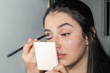 face of a latin woman applying makeup to her eyelids with a small brush while looking in the mirrorの写真素材