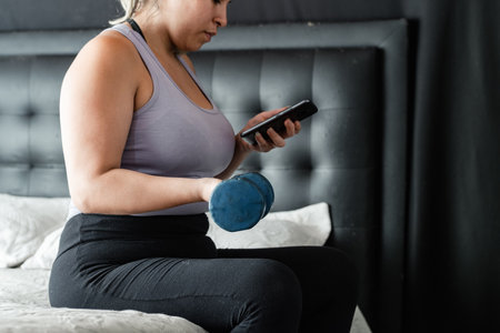 young latina woman sitting on her bed looking at her cell phone while exercising with her blue dumbbellの写真素材