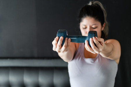 young latina woman very concentrated lifting a blue dumbbell, working her muscle endurance.の写真素材