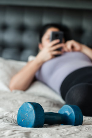 close-up of blue dumbbell on bed, in the background a young woman using her cell phone lying on the bedの写真素材