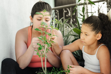 little brown-skinned girl next to her mother looking curiously at the leaves of the green garden plantの写真素材