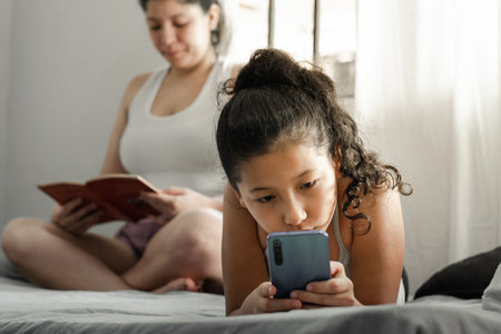 little girl looking curiously at her cell phone, lying on the bed while her mother reads a book.の写真素材