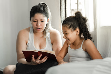 brown girl smiling while her mother reads a book to her, sitting on top of the bedの写真素材