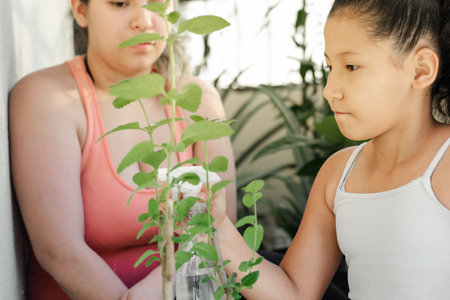 little brown-skinned latina girl sprinkling water on the green silver, while her mother looks on.の写真素材